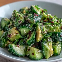A vibrant bowl of smashed cucumber and avocado salad, drizzled with sesame dressing and topped with green onions and sesame seeds.